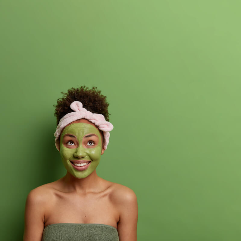 spa-beauty-treatment-skin-care-concept-positive-female-applies-face-peeling-mask-stays-young-beautiful-concentrated-with-broad-smiles-wears-headband-poses-green-wall-copy-space (1)